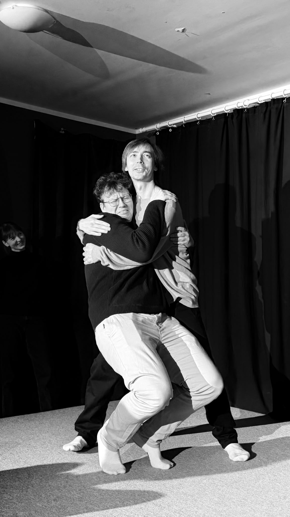 Two performers locked in a dramatic embrace on stage, in black and white